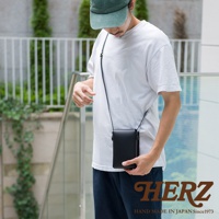 一名男子身著白色T恤和深藍色長褲，斜挎一個黑色皮革手機包。手機包為長方形，以細長黑色皮革背帶連接，包蓋處有磁扣設計，整體為簡約風格。