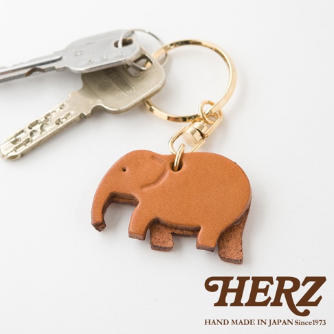 皮革製成棕色大象造型鑰匙圈，搭配金色鑰匙圈和掛鉤，與銀色鑰匙一同置於白色背景上。