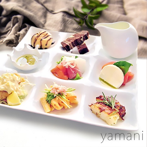 白色陶瓷九格小菜盤，盛有各式精緻小點，包含：裝飾有焦糖醬的甜點、巧克力大理石紋蛋糕、搭配醬料的白色小碗、生魚片、番茄與莫札瑞拉起司、沙拉、竹筍料理，以及烤魚塊，盤旁另有一白色小湯盅。