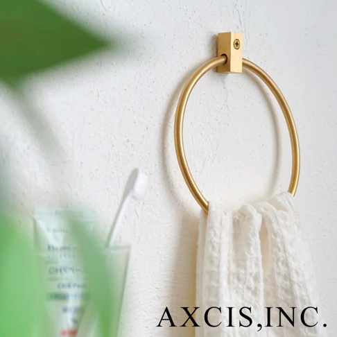 黃銅材質的圓形毛巾環，以簡潔的方形底座固定於白色紋理牆面上，環上懸掛一條白色格紋毛巾，左側為模糊的牙刷與牙膏。