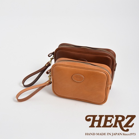 兩款職人手作皮革萬用手提包，一款為淺棕色，另一款為深棕色，皆有可拆式扣環手腕帶，以簡潔的設計和精緻的縫線呈現。