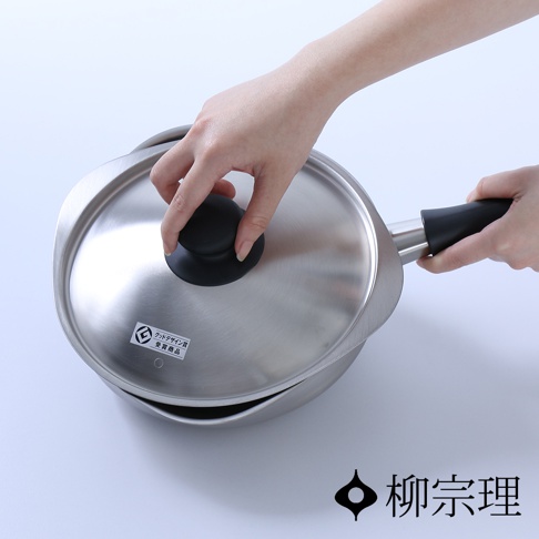 一雙手正持握柳宗理霧面不鏽鋼單手鍋，上方視角展示其獨特的流線型鍋蓋與黑色耐熱把手設計，鍋蓋上貼有象徵獲得優良設計獎的標籤，整體呈現簡約精緻的金屬質感。