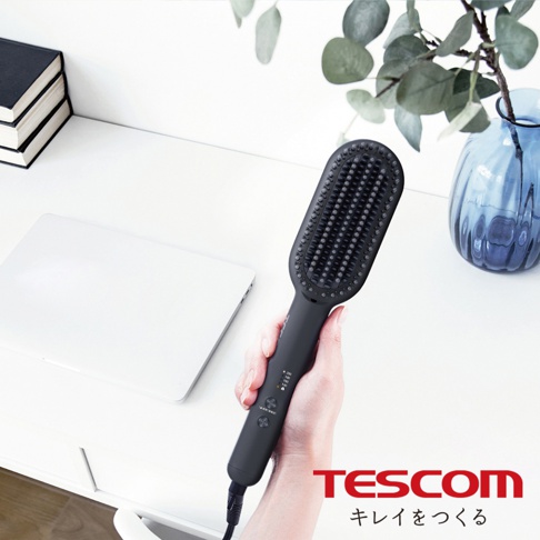 手持黑色負離子直髮造型梳，梳齒密集，手柄處有溫度顯示與按鈕，背景為白色桌面，擺有筆記型電腦、書籍及藍色花瓶。