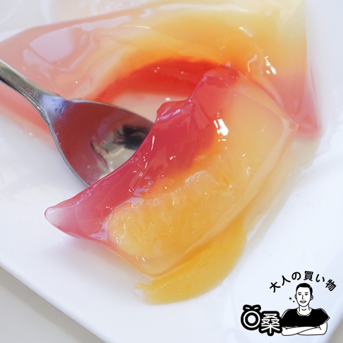一塊三角形的日本國產水果果凍，呈現粉紅色與橘黃色漸層，果凍內含切塊水果，搭配金屬湯匙，置於白色盤子上。