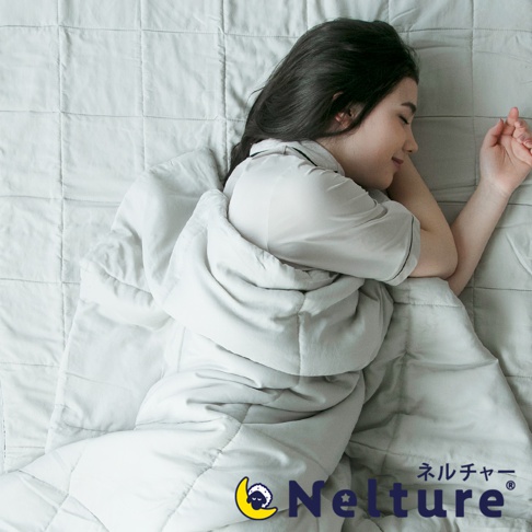 一位女性蓋著一條淺灰色的Nemulia五層散熱涼感單人被，被子質地輕薄，表面有細緻的格紋縫線。