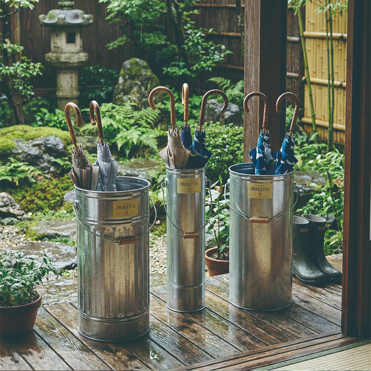 日式庭院裡，雨水滴落，三個金屬傘桶整齊擺放在木質地板上，裡面各插著幾把帶有木質彎把的雨傘，傘桶上印有 OBAKETSU 字樣，背景是茂密的綠色植物和竹林。