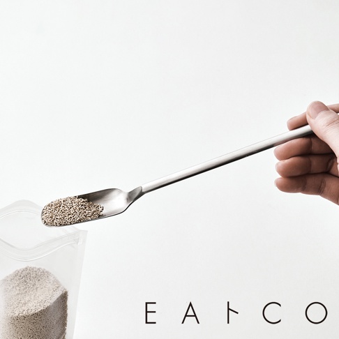 一只手正拿著一把日製金屬製的小長勺，勺頭裝有細小的顆粒狀物體，旁邊有一個裝有相同顆粒的透明塑膠袋。