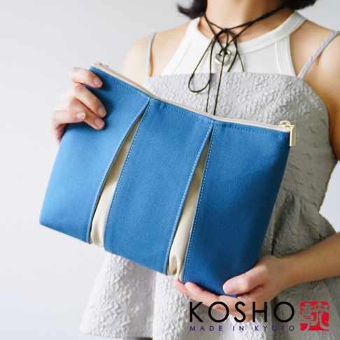 一位女性雙手拿著一個京都KOSHO品牌的ougi帆布化妝收納包，該收納包由藍色與米色帆布拼接而成，設計簡約且具有立體感，右下角印有KOSHO MADE IN KYOTO的品牌標誌。