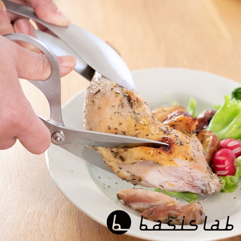 一只手正使用一把銀色不鏽鋼料理剪刀，剪切盤中一塊烤雞。烤雞表面有香料，盤中還有生菜和紅蘿蔔。