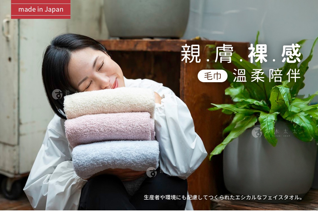 一位年輕女性閉眼微笑，懷抱著三條堆疊在一起、色彩柔和的有機棉毛巾，分別為米色、粉色與淺藍色。畫面左上方標註日本製造，右側標題強調親膚裸感與溫柔陪伴，下方說明這是一款兼顧生產者與環境的友善毛巾。整體氛圍舒適溫暖，呈現日本津OBORO有機棉毛巾的高品質質感。