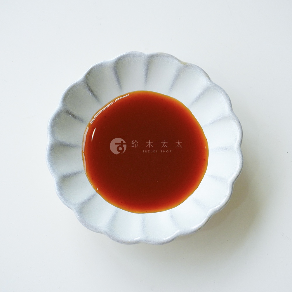 一個白色花瓣造型的陶瓷碟子，正中間盛裝著深琥珀色的朝紫大蒜酸橘醋風味萬用調味醬，醬汁質地呈現清澈流體狀，背景為簡約乾淨的純白色。