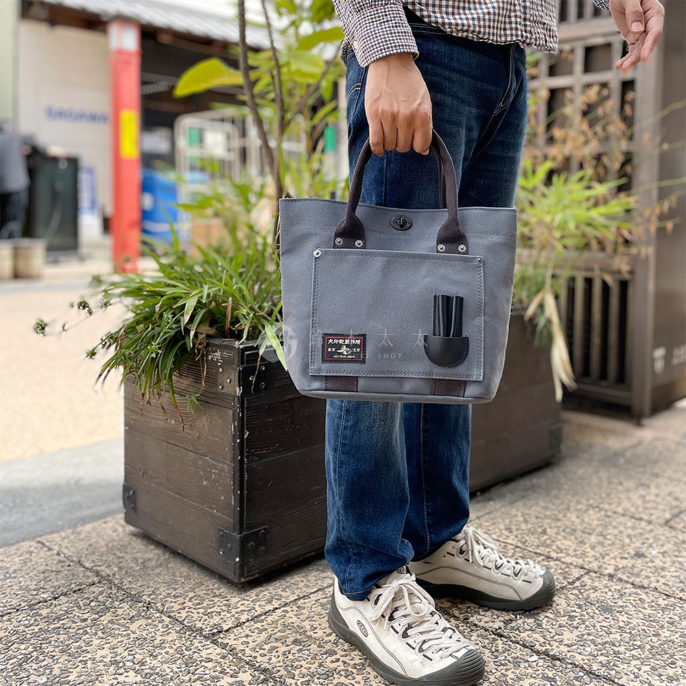 灰色帆布製成的水桶托特包，提把為深棕色皮革，包包正面有一個方形外袋，內有黑色筆狀物。