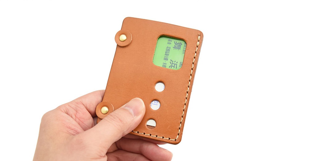 一手拿著日本HERZ品牌手工皮革交通卡收納夾KC-103，卡夾為焦糖棕色，側面縫線精緻，上方有一顆鉚釘，卡夾內可見一張綠色日文卡片，卡夾前方有三個圓孔設計。