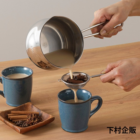 一只手正傾倒不鏽鋼牛奶鍋中的奶液，通過濾網過濾肉桂棒和香料，倒入藍色陶瓷馬克杯中。