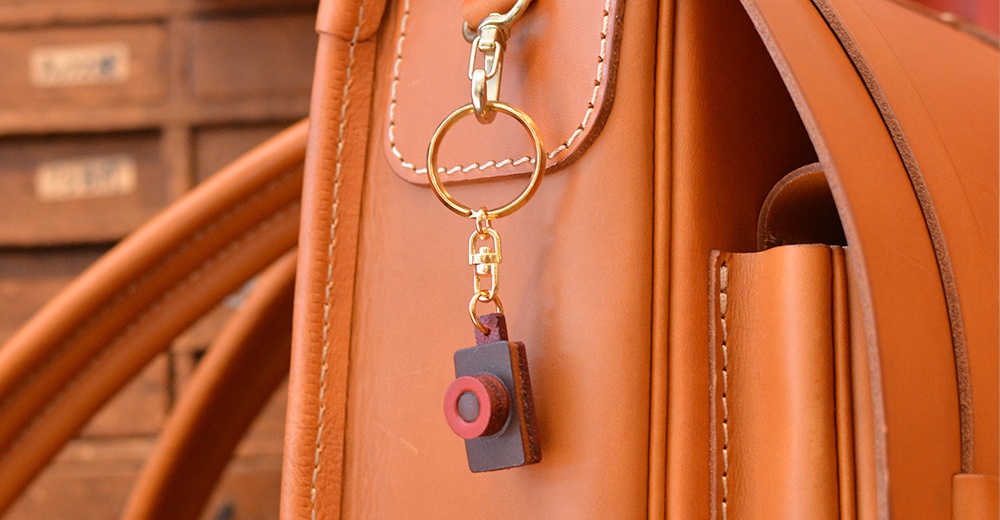 職人手作皮革相機造型鑰匙圈，以棕色皮革製成，搭配紅色鏡頭裝飾，掛於同色系皮革包上，配有金色鑰匙圈與扣環。