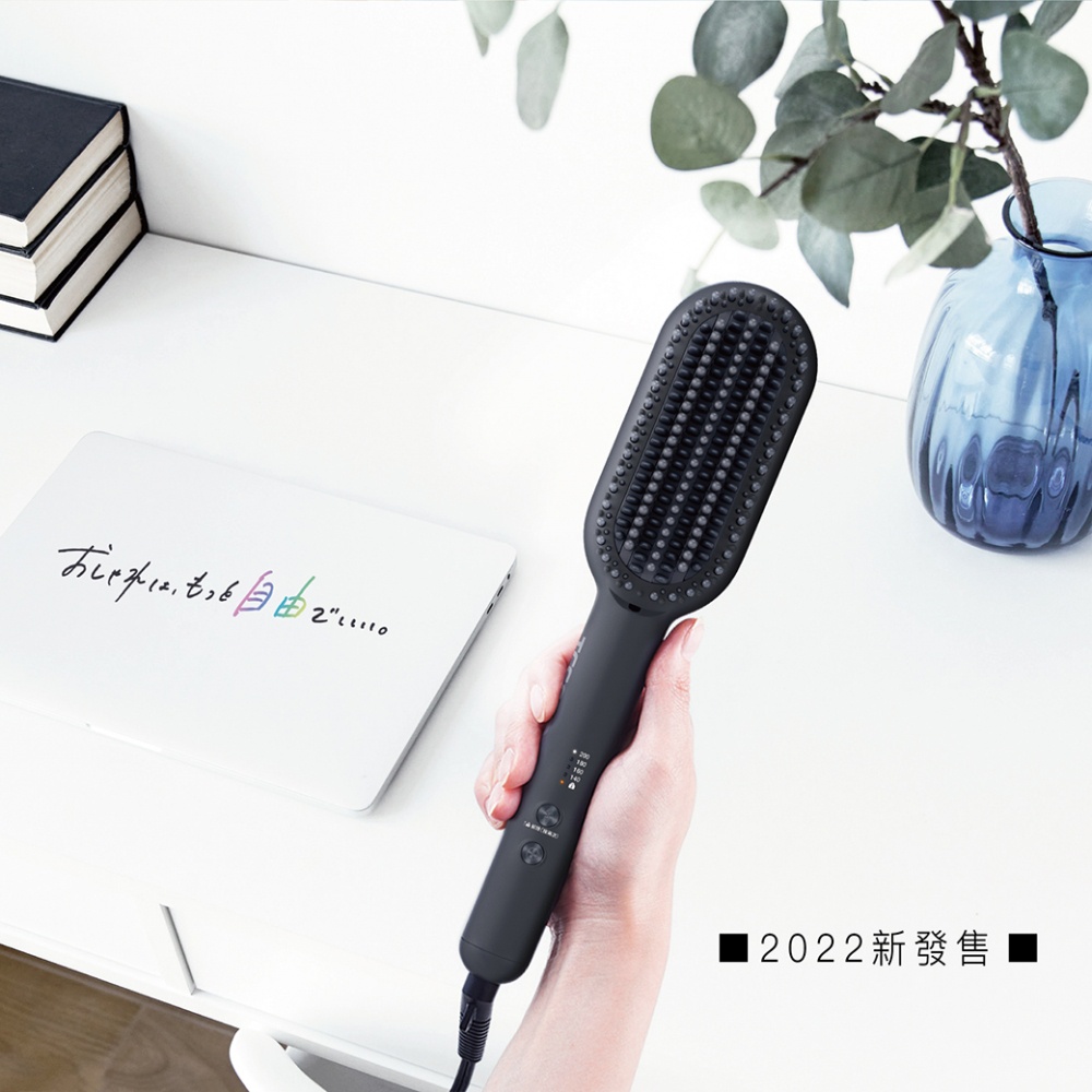 一只手正握著一支黑色的負離子直髮梳，梳子上方有許多細密的梳齒，梳柄上顯示有溫度設定的指示燈。背景是一個淺色的桌面，上面放著一本筆記本電腦、幾本書和一個藍色花瓶。