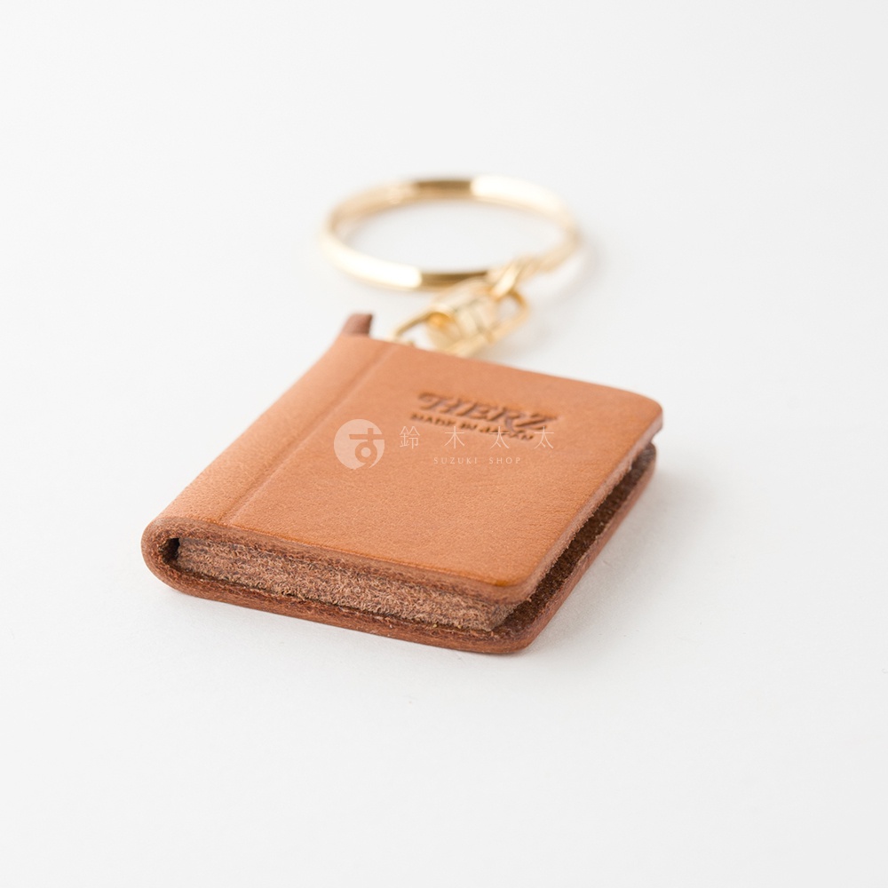 職人手作皮革書本造型鑰匙圈，採用淺棕色皮革製成，搭配金色鑰匙圈，質感細緻，封面壓印有品牌標誌。