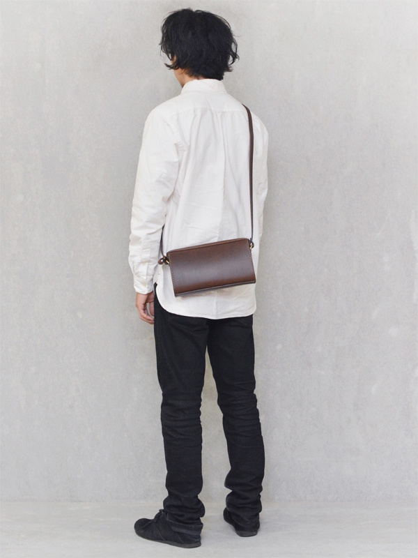 一位男士身穿白色襯衫和黑色長褲，側背一個深棕色皮革拉鍊單肩包。