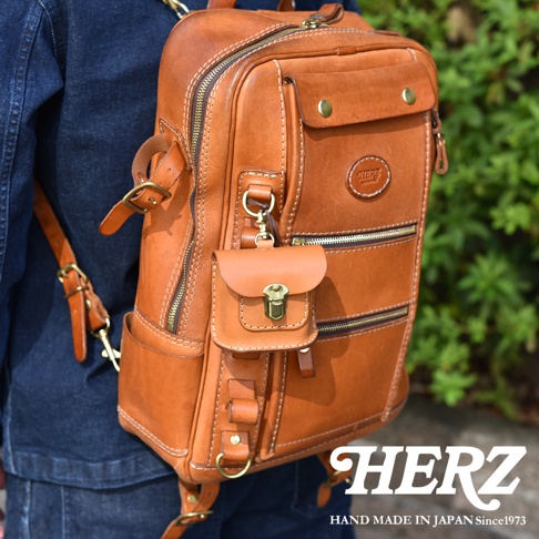駝色皮革後背包，配有小巧的皮革掛飾，可收納耳機。包款採用精緻車縫線，有多個拉鍊口袋及金屬扣環，展現職人手作質感。