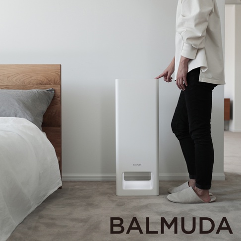 The Pure 空氣清淨機，白色直立式機身，設有簡潔的進氣口，位於臥室地板上，旁邊是木製床頭和床鋪，一位身著白色上衣、黑色長褲的女士正伸手觸碰機身。