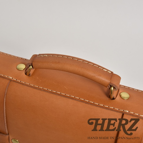 職人手作皮革傳統歐風三用學生包，展示包款的皮革手把與金屬卯釘細節，淺棕色皮革搭配米白色車線，質感細緻。