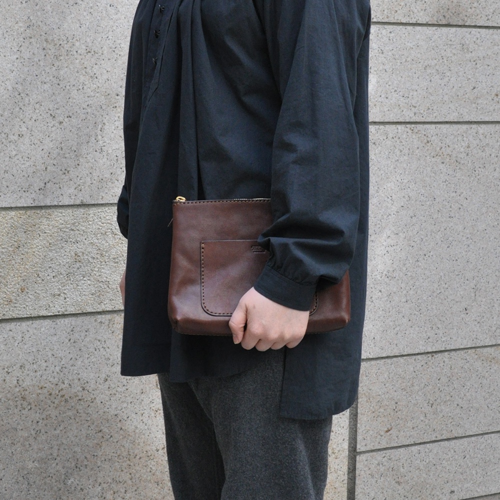 一件咖啡色皮革手拿包，有外部口袋和拉鍊開合，由手持。