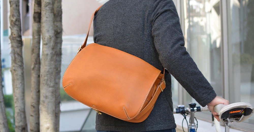 側背著一個棕褐色皮革翻蓋郵差包，包包有著復古風格的縫線和金屬扣環，搭配灰色夾克。