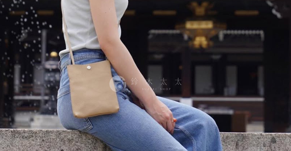 淺褐色皮革材質的簡約隨身手機包，以斜背方式由女性展示，包款設計簡潔，附有銀色金屬按扣。