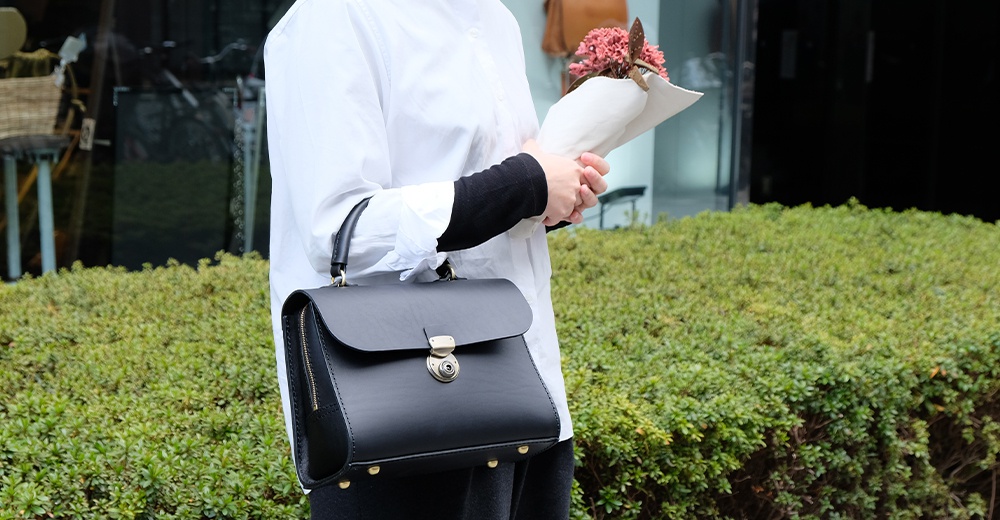 日本HERZ職人手作黑色皮革多功能收納單肩包，配有金屬鎖扣和手提把，手持一束粉色小花。