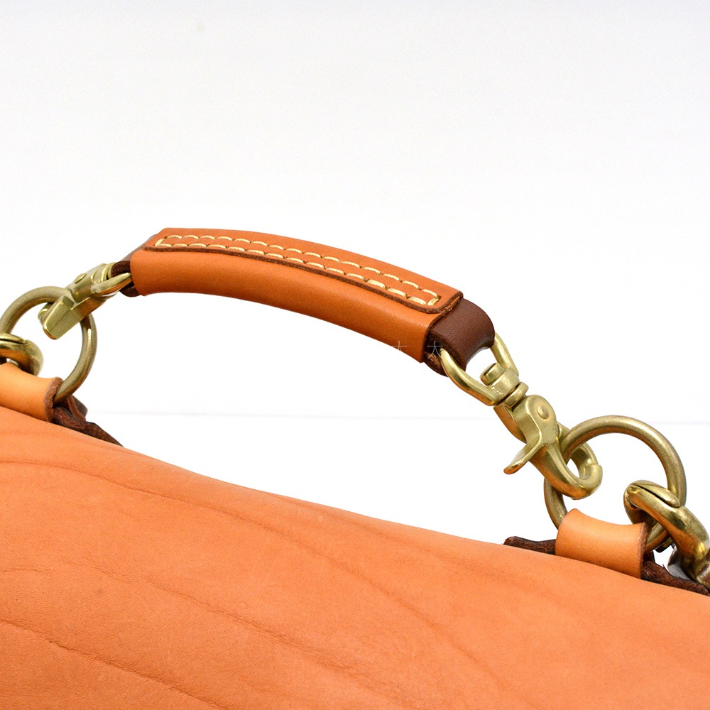 淺棕色皮革郵差包，帶有對比色的棕色皮革提把，提把以白色縫線裝飾，並連接至黃銅色金屬掛鉤和圓環。
