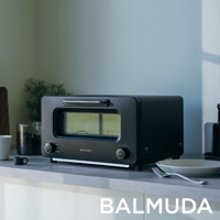 黑色 BALMUDA The Toaster Pro 蒸氣烤麵包機 K11C-SE，配有銀色把手和兩個旋鈕，擺放在檯面上，旁邊有咖啡杯、小碟子和叉子。