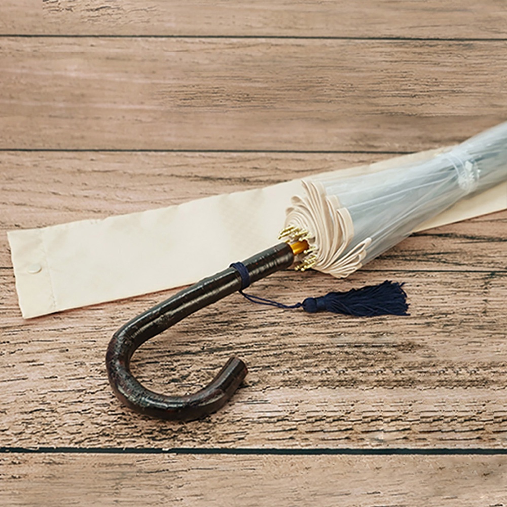 職人級樺木手柄透明傘，傘骨與傘布為淺米色，傘面為透明材質，傘柄處繫有深藍色流蘇，附有同色系收納袋。