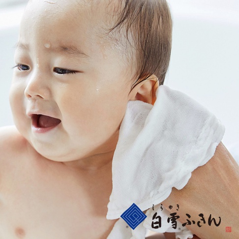 一張嬰兒的臉部特寫，左側是嬰兒的臉，右側是一隻手拿著一條白色、紋理清晰的洗澡巾，正在擦拭嬰兒的臉頰。