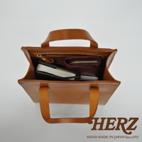 職人手作皮革直式托特包 T-44 XS，以淺棕色皮革製成，包內可見筆記本、手機及紅色零錢包，頂部有拉鍊開合及提把，整體風格簡約。