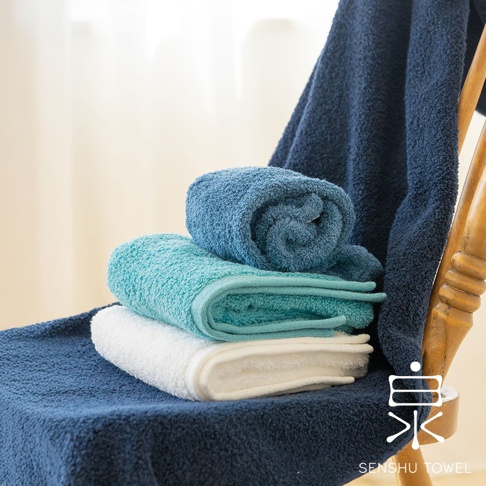 堆疊著三條迷你浴巾，顏色分別為深藍、淺藍和白色。浴巾材質為長纖維，質地柔軟蓬鬆。其中一條淺藍色浴巾捲起，置於其他浴巾上方。背景為木椅與柔和的光線。