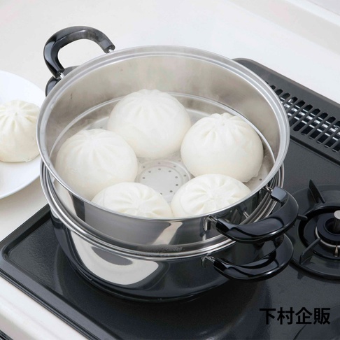 不鏽鋼雙層蒸氣料理鍋，鍋內裝有數個圓潤飽滿的白色包子，蒸氣從鍋中冒出。鍋身帶有兩個黑色隔熱手柄，置於黑色爐灶上。
