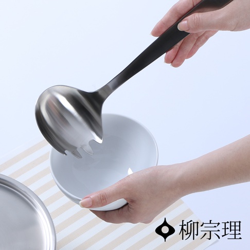 一只手正持握著不鏽鋼叉杓，叉杓頭部有四齒設計，用於撈取食物。叉杓的柄部為黑色。下方有一個白色碗，碗旁有條紋墊。