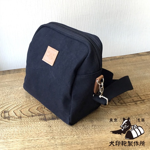 深藍色石洗帆布散步包，方正立體造型，正面有一個淺棕色皮革方形標籤，上面有可愛的狗狗圖案壓印。包包配有黑色織帶和金屬扣環。右下角有日文圖案標誌，內容為「東京 淺草 犬印鞄製作所」。