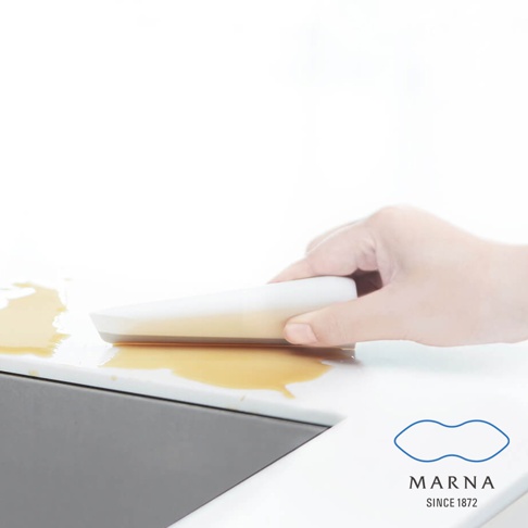 白色廚房清潔刮板，搭配一隻手正在使用它來清除檯面上淺褐色液體。刮板採用簡潔的流線型設計，材質看似塑膠或矽膠。