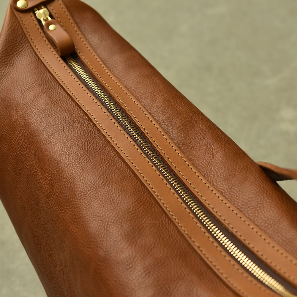 職人手作皮革Organ單肩半月包，採用鞣製皮革製成，具有溫潤的棕色調。包身呈現半月弧形，並以精緻的黃銅色拉鍊和縫線裝飾，材質觸感細膩。