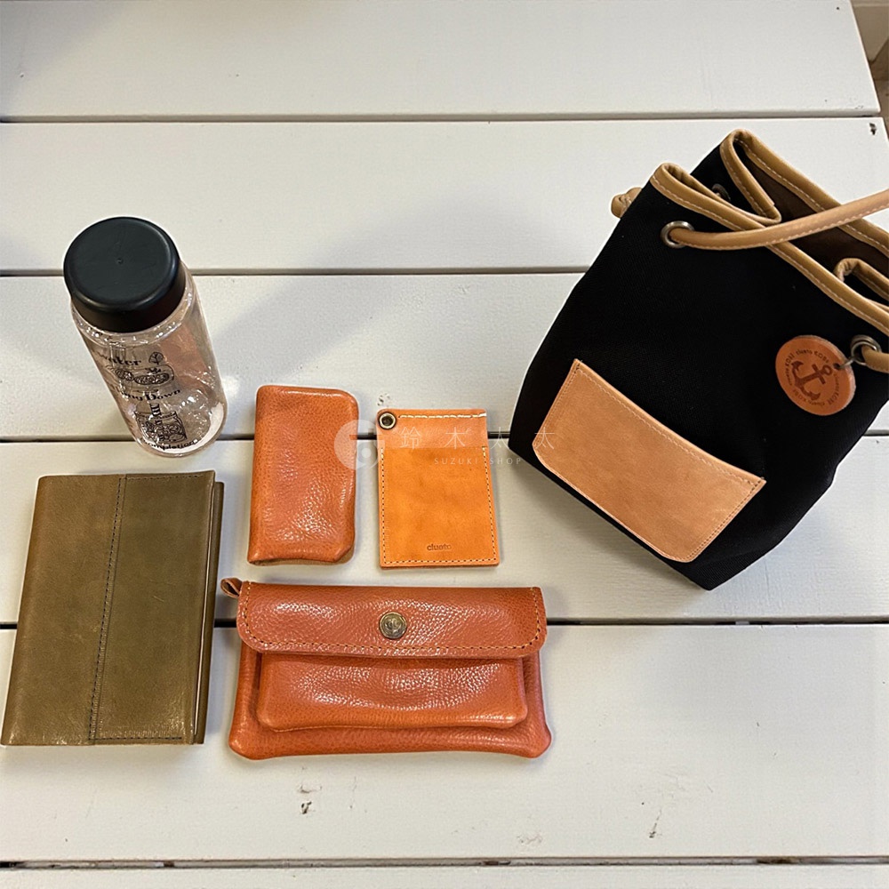 圖中有一個黑色束口包，搭配橘色和棕色的皮件，以及一個透明水瓶和橘色的皮夾。