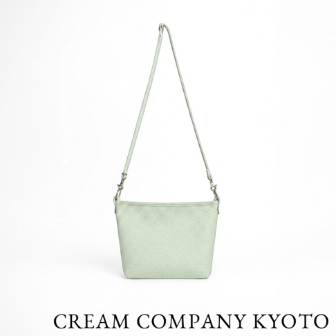 這款由 CREAM COMPANY KYOTO 出品的薄荷綠色牛皮側背包，包身採用柔軟耐用的皮革材質製成，呈現簡約的梯形輪廓與細膩的荔枝紋理。包包配有可拆卸的細肩帶，下方印有品牌名稱字樣，整體設計簡潔輕便，展現日系職人手作工藝。