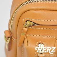 職人手作皮革單肩包，使用淺棕色皮革，搭配金色拉鍊和金屬配件，縫線明顯，質感良好，風格簡約。