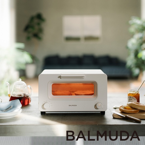 白色 BALMUDA 蒸氣烤麵包機，設有玻璃門，內部有兩片正在烘烤的麵包，底部有旋鈕和按鈕，表面光滑。