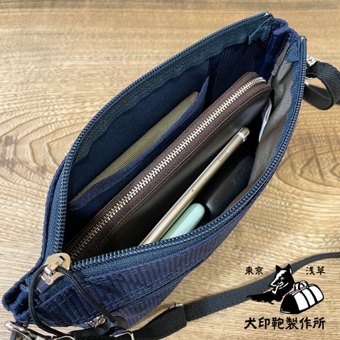 深藍色刺子織斜背包，拉鍊敞開，露出內部收納空間，可見錢包、手機、薄型手機殼等物品。