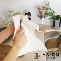 一雙手正在使用白色絎縫的廚房拭巾擦拭餐具，拭巾邊緣有灰色滾邊。背景為廚房水槽、咖啡濾杯與綠色植物。