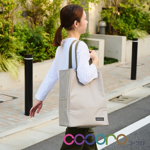 一位女性提著一個米色帆布材質的2WAY手提收納折疊推車，推車上有綠色提把，包身側面有兩個口袋，並印有品牌Logo。