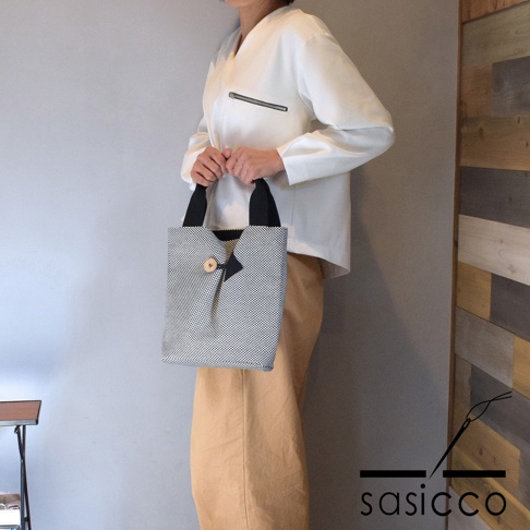 一位女士手持一個直式收納托特包，包身為黑白細格子圖案的刺子織布料，配有黑色提把，正面有一個木質鈕扣和一個黑色三角形裝飾。