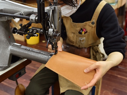 職人正在使用工業縫紉機縫製一個淺棕色皮革長夾，長夾表面光滑，邊緣有整齊的縫線，顯示出精湛的手工藝。