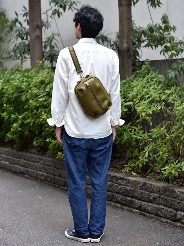橄欖綠皮革立體運動斜背包，側背於身著白色襯衫與牛仔褲的男性身上，包款設有拉鍊開合與可調節背帶。